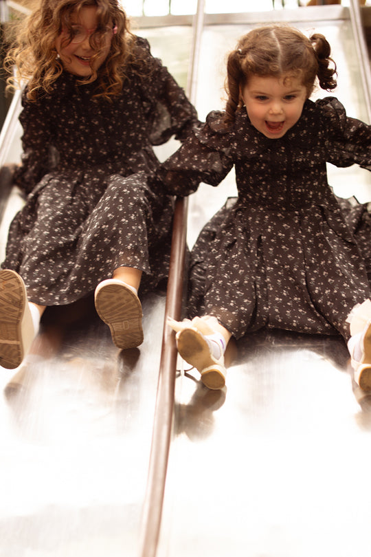 Two young girls, dressed in matching Ivy City Co Mini Sophia Long Sleeve Dresses in Noir Ditsy Chiffon, slide side by side down a shiny playground slide, smiling and having fun.