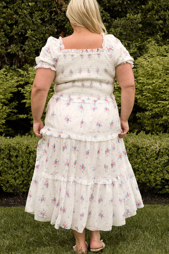 A woman with blonde hair stands outdoors, facing away, wearing the Ivy City Co Emily Dress in Heritage Soft Ditsy Floral—a white, floral print midi dress featuring puffed short sleeves and a tiered skirt. Barefoot on the grass and surrounded by green bushes, she perfectly showcases this nursing-friendly maternity dress.