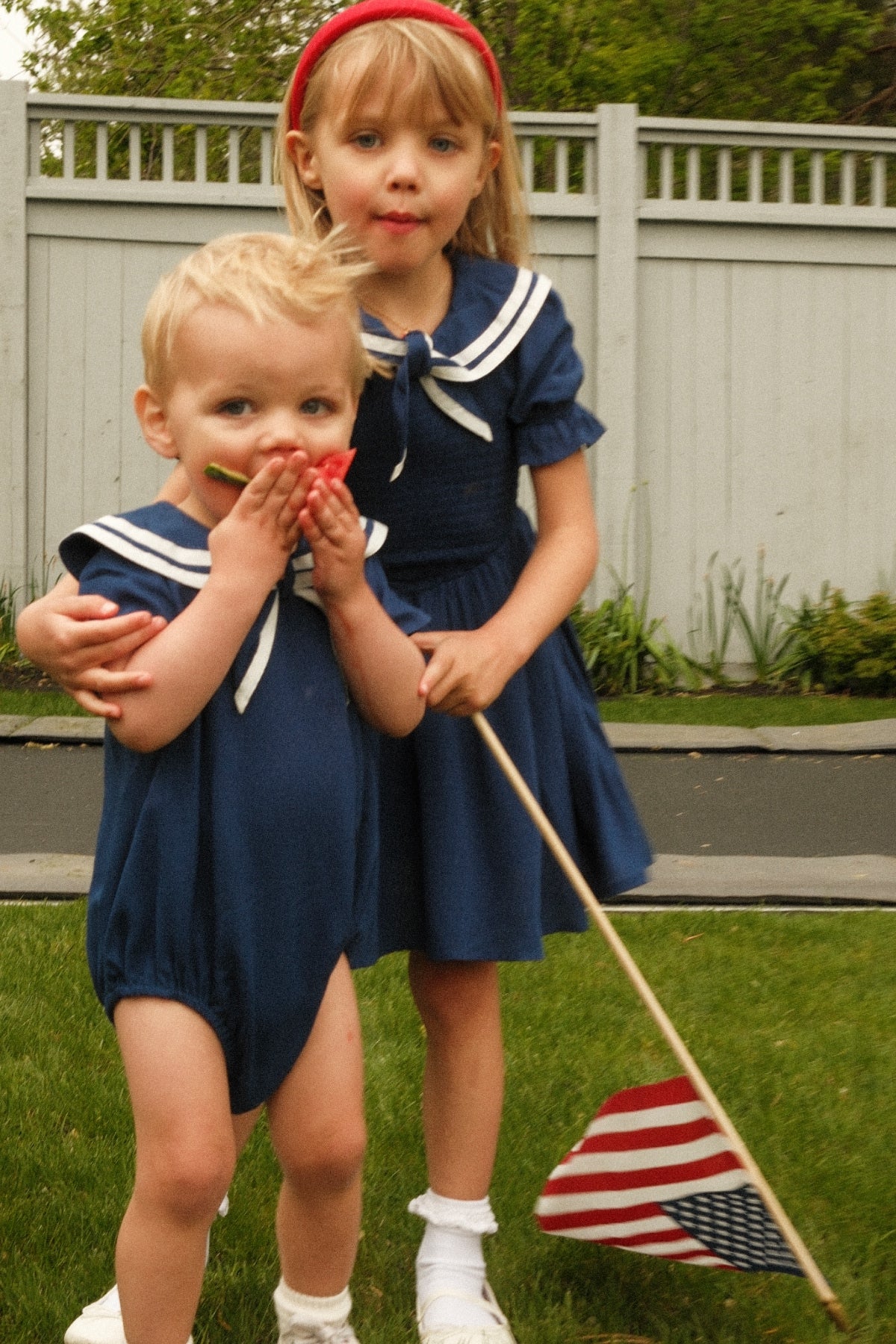 Two young children in blue sailor outfits, including the Mini Alice Dress in Navy by Ivy City Co, stand on grass. The older child holds a U.S. flag and wears a red headband, while the younger child enjoys a strawberry. A gray fence and lush greenery are visible in the background, creating a delightful Mom and Me matching moment that’s perfect for summer celebrations.