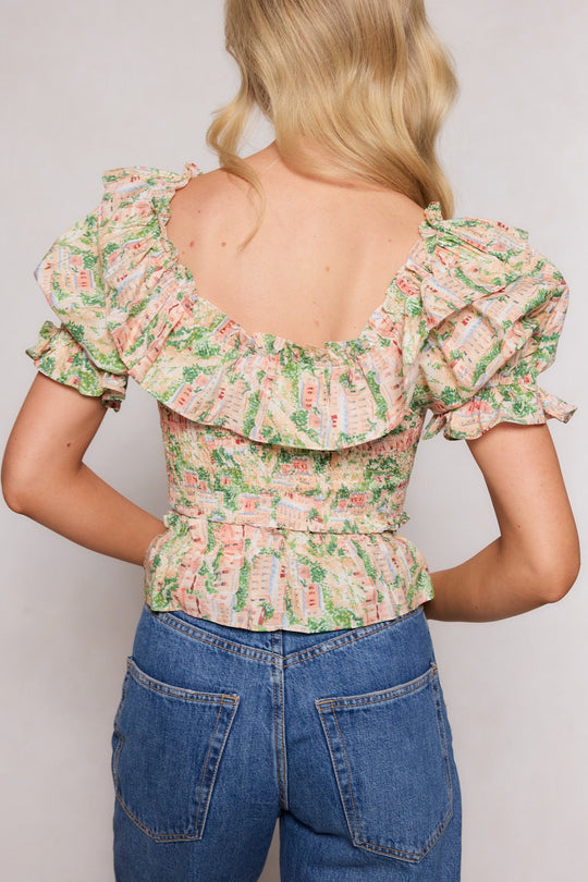 A woman with wavy blonde hair is seen from the back against a neutral background, wearing Ivy City Co’s Julian Top in Cream—a floral ruffled blouse with puff sleeves—paired with high-waisted blue jeans.