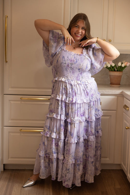 A woman wearing the Winnie Dress in Lavender Aquarelle Fleur by Ivy City Co poses in a kitchen, smiling with her hands framing her face. She stands near cream cabinets and a pot of tulips on the counter.