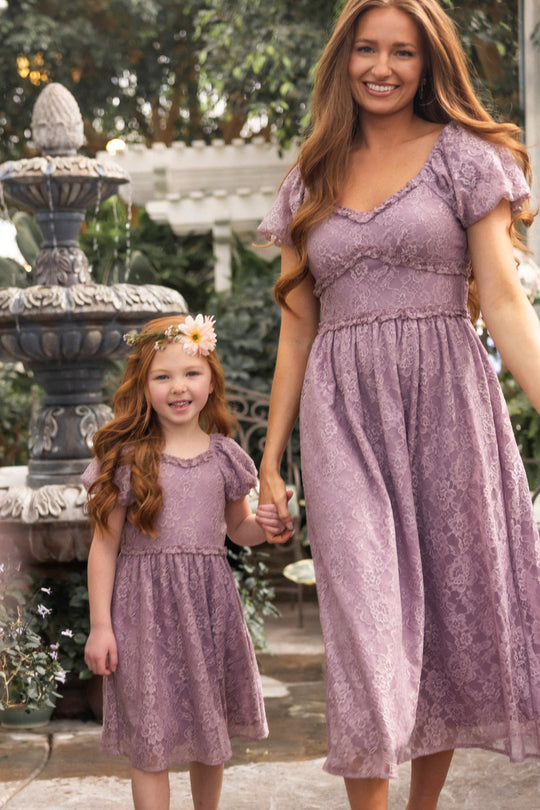 A woman and a young girl, both with long red hair, wear matching Ivy City Co Kiara Dresses in Hyssop Lace. They are holding hands and smiling, standing in front of a garden fountain surrounded by greenery and flowers.