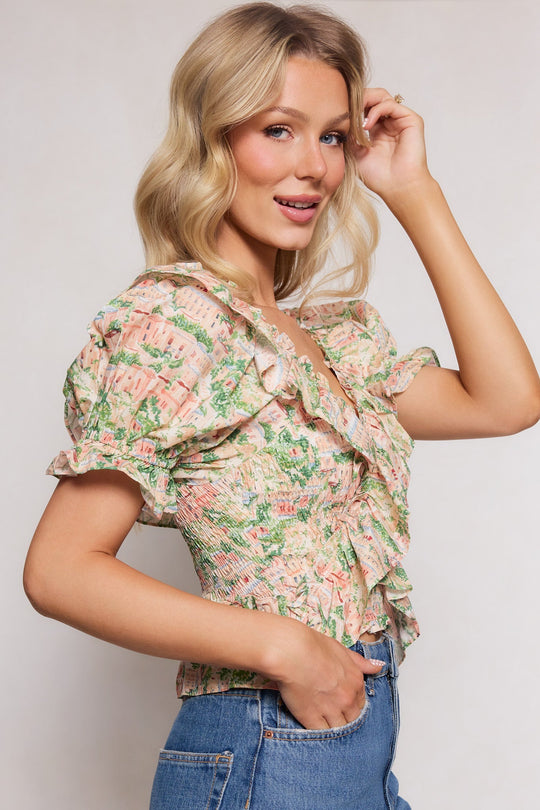 A woman with wavy blonde hair stands in profile against a neutral background, smiling and lightly touching her hair as she wears the Ivy City Co Julian Top in Cream paired with high-waisted blue jeans.