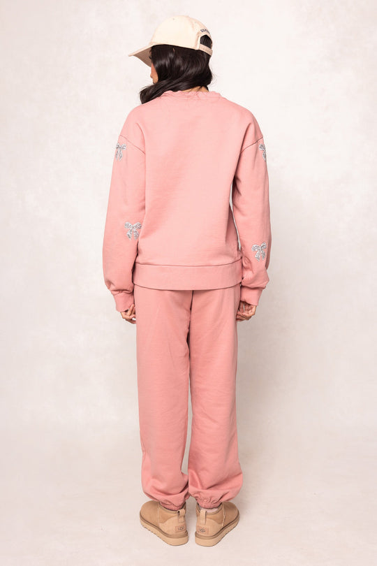 A person stands facing away, wearing the Ivy City Co Micah Crew Neck Sweatshirt in Pink & Bows with matching sweatpants featuring subtle embroidered details on the sleeves. They’ve paired the outfit with tan sneakers and a beige baseball cap, set against a plain light background.