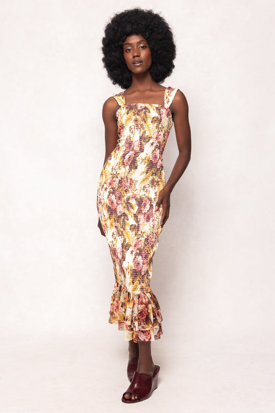 A woman with curly hair wears the Ivy City Co Mira Dress in Garden Rose—a sleeveless, floral-print midi dress with a ruffled hem—and pairs it with burgundy open-toe shoes, standing against a plain light background.