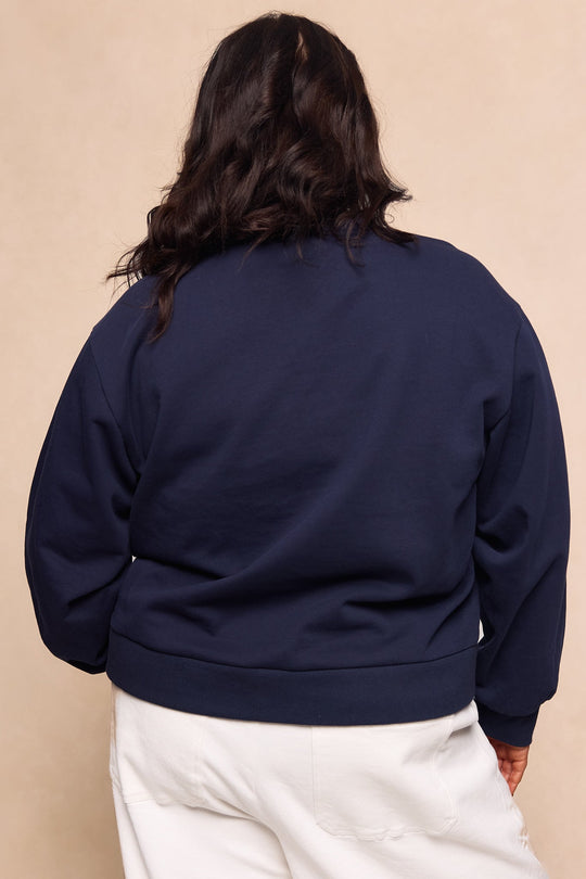 A person with long dark hair, wearing the Ivy City Co Kinsley Sweatshirt in Navy and white pants, stands with their back to the camera against a beige background.