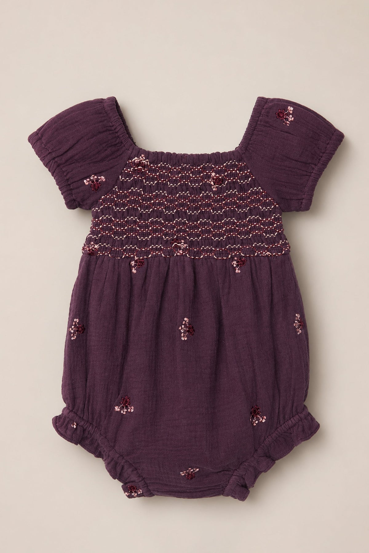 The Baby Acantha Romper in Aubergine Embroidered Gauze by Ivy City Co is a purple cotton romper featuring short puff sleeves, ruffled leg openings, a smocked chest, and delicate embroidered floral patterns scattered across the fabric.