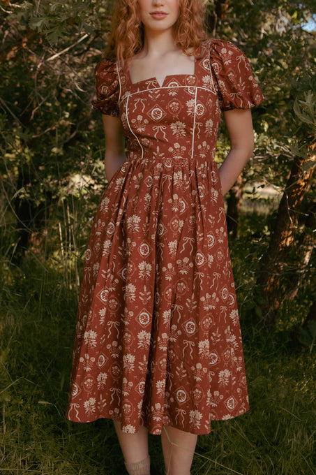 A woman with wavy red hair wears the Nash Dress in Sepia Bows by Ivy City Co, a rust-colored, cottagecore midi dress with puff sleeves and a white bow print, standing outdoors among green trees and grass. Her face is partially out of frame.