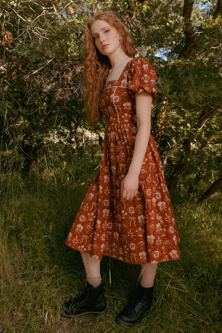 A person with long wavy red hair, wearing the Ivy City Co Nash Dress in Sepia Bows and black boots, stands outdoors on grass among leafy trees and sunlight.