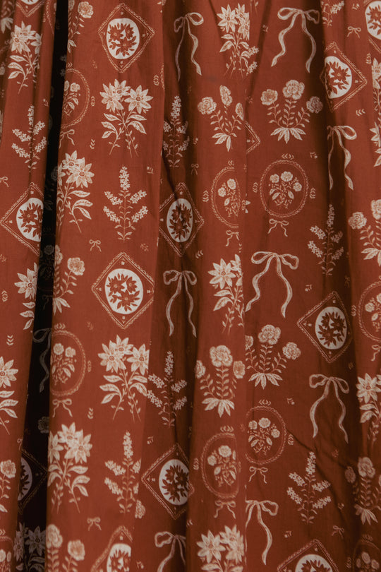 The Nash Dress in Sepia Bows by Ivy City Co features red fabric with white floral and ribbon patterns, adorned with circular and diamond motifs—ideal for a cottagecore-inspired or elegant puff sleeve look.