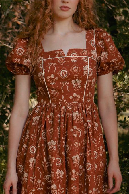 A woman with long wavy hair wears the Ivy City Co Nash Dress in Sepia Bows—a rust-toned cottagecore midi dress with puffed sleeves, square neckline, white piping, and bow print—standing outdoors amid green foliage.