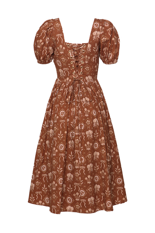 The Nash Dress in Sepia Bows by Ivy City Co is a brown cottagecore midi dress with puffed sleeves, square neckline, lace-up corset back, flared skirt, and a charming white floral and ribbon pattern.