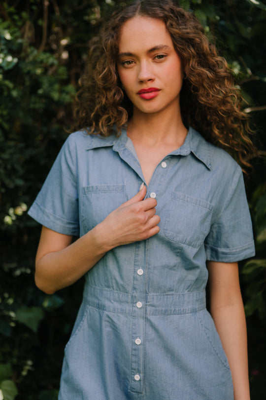 A woman with curly hair stands outdoors before green foliage, wearing the nursing-friendly Natalia Romper in Chambray by Ivy City Co, and looks at the camera with a neutral expression.
