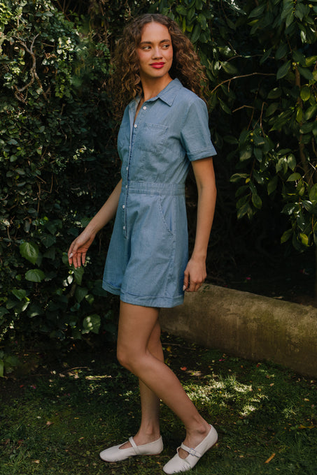 A woman with curly hair wears the Ivy City Co Natalia Romper in Chambray and white shoes as she stands on grass, surrounded by green foliage and trees.