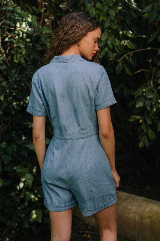 A woman with wavy hair stands outdoors among green foliage, facing away from the camera, wearing the Ivy City Co Natalia Romper in Chambray.