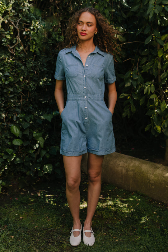 A woman stands on grass in the Ivy City Co Natalia Romper in Chambray—short-sleeved, button-up, with pockets. With curly hair and red lipstick, she’s surrounded by lush greenery. This nursing-friendly romper is perfect for casual days.