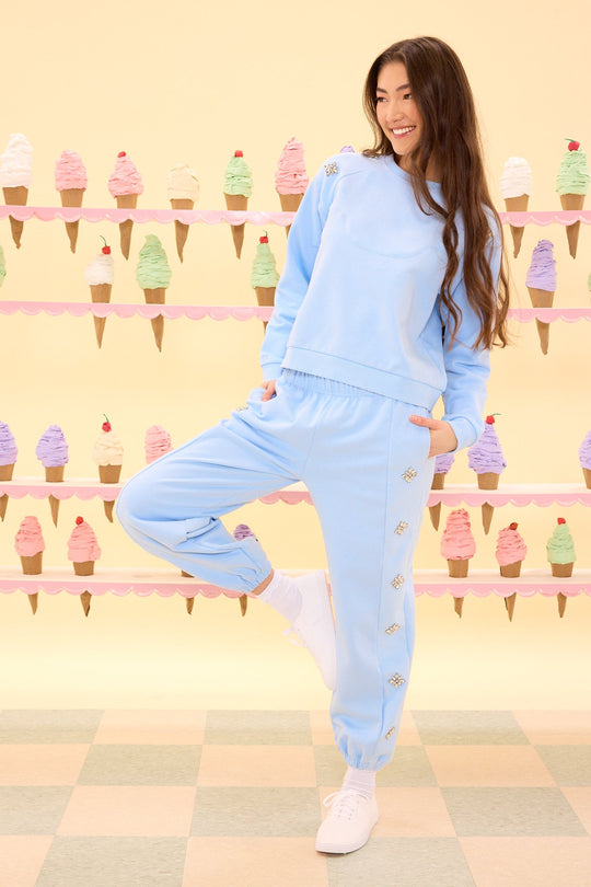 Wearing the Casey Crewneck Sweatshirt in Light Blue by Ivy City Co and white sneakers, a woman poses with one leg up, smiling in front of a pastel wall adorned with colorful ice cream cones. Her outfit shimmers subtly with crystal details.