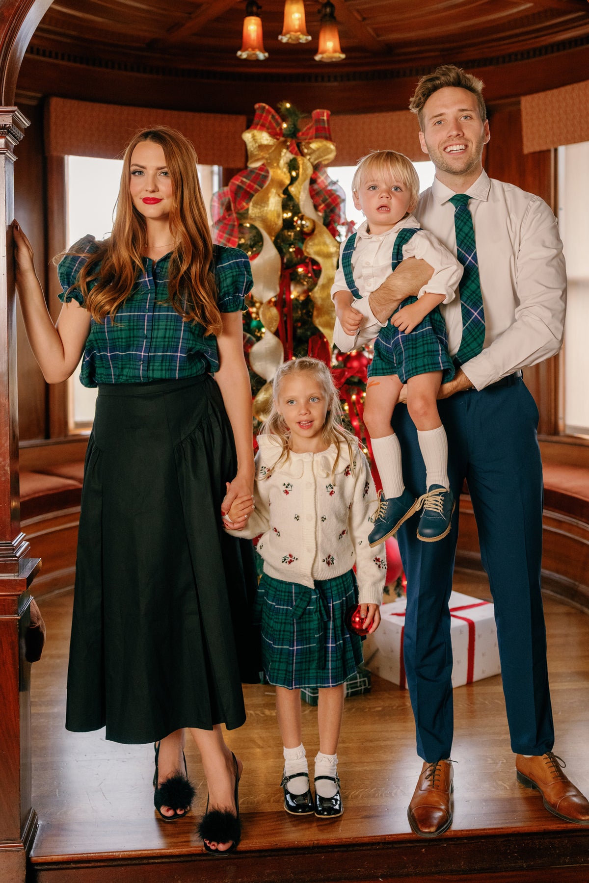 A family of four poses indoors by a Christmas tree, dressed in coordinated green plaid outfits. The two young children wear the Ivy City Co Nicolette Blouse in Emerald Plaid, featuring Peter Pan collars, as they stand between their parents. Festive decorations and wrapped gifts add to the holiday scene in the background.