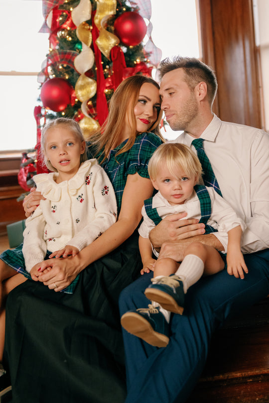 A family of four sits together in front of a decorated Christmas tree, smiling and dressed in festive outfits. The youngest daughter beams in her Ivy City Co Nicolette Blouse in Emerald Plaid, its charming Peter Pan collar adding an extra touch of holiday cheer to the joyful scene.