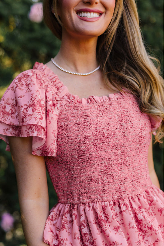 A woman smiles outdoors amidst green foliage, wearing the Ivy City Co Grace Dress in Desert Heritage Rose—a pink floral maxi dress with fluttering ruffled sleeves that’s maternity friendly and elegantly paired with a pearl necklace, offering comfort and style for any occasion.