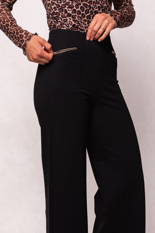 A woman wearing the Alyssa Pop Ponte Pants in Black by Ivy City Co, featuring a slimming fit, tummy control, and a convenient zipper pocket, pairs them with a long-sleeved leopard print top as she holds the zipper with her right hand. Set against a plain light-colored background, these versatile pants are the perfect day-to-night staple.