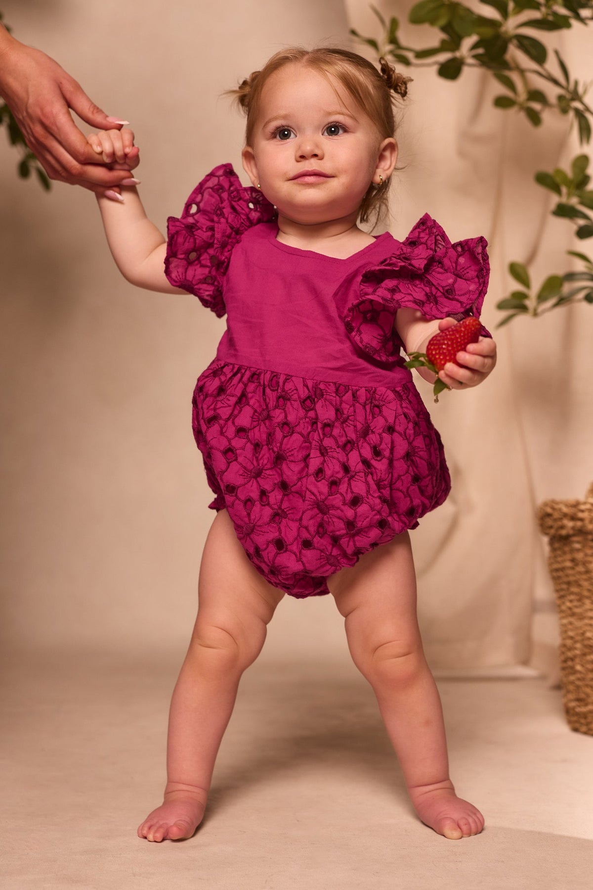A toddler wearing the Ivy City Co Baby Hattie Romper in Magenta, featuring delicate floral embroidery on soft cotton, stands indoors near a leafy plant, holding a strawberry in one hand and an adult’s hand with the other. This charming romper makes a perfect Easter outfit.