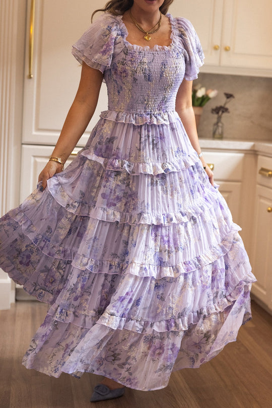 In a sunlit kitchen, a woman wears the Ivy City Co Winnie Dress in Lavender Aquarelle Fleur, its flowing layered chiffon, puffed short sleeves, and delicate ruffles highlighted as she holds out the skirt to reveal its romantic florals and breathtaking fullness—a dreamy choice for a spring wedding.