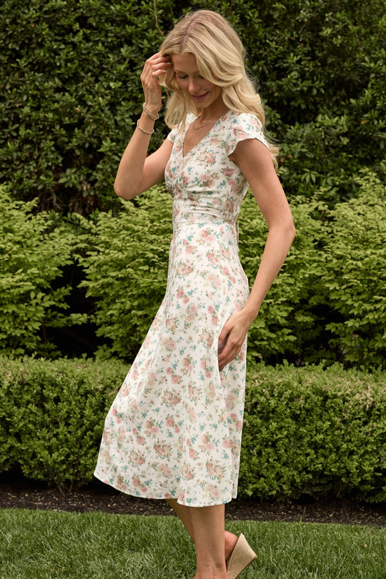 A woman wearing the Ivy City Co Fiona Dress in Countryside Peony Blossom stands on grass, smiling and touching her hair, with green bushes and trees in the background.