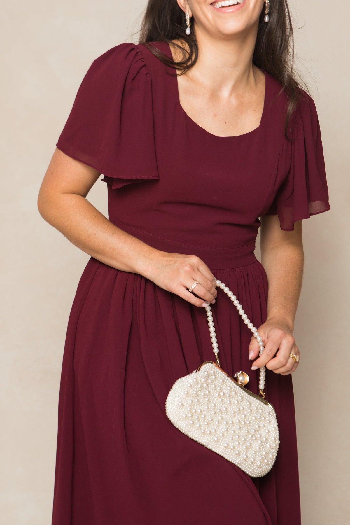 A woman smiles while wearing the Ivy City Co Abbie Dress in Wine Chiffon and holding a white beaded handbag. Her dark hair is down, and she accessorizes with pearl earrings against a neutral background.