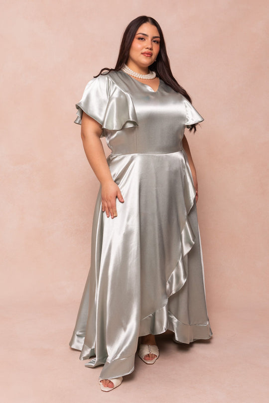 A woman models the Callie Dress in Sage Satin by Ivy City Co—a lustrous sage green gown featuring ruffled sleeves and a flowing hem—styled with a pearl choker necklace and open-toe heels, as she poses elegantly against a soft beige backdrop.