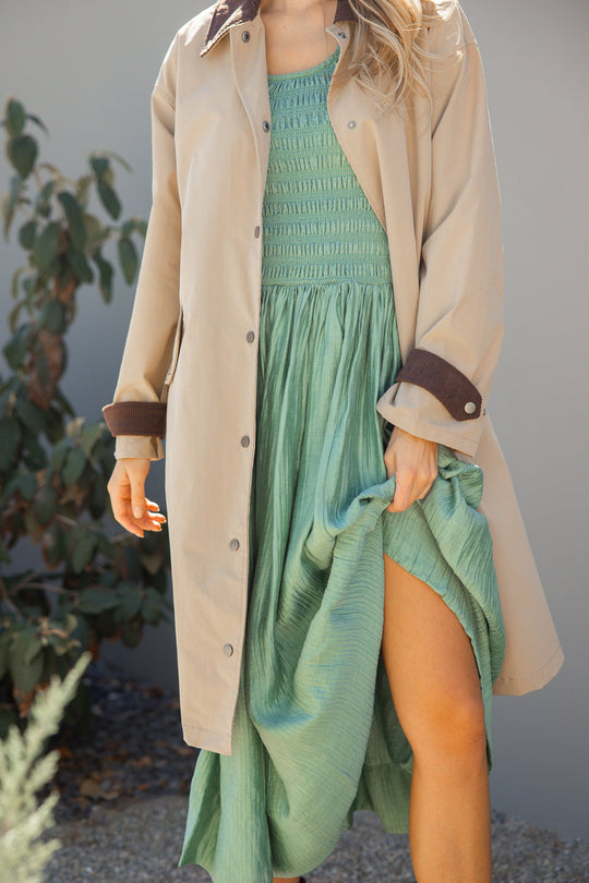 A woman stands outdoors among some plants, wearing the Ivy City Co Marlow Dress in Green—a light green textured midi dress with a smocked bodice—paired with a beige trench coat. She lifts the side of her dress to reveal her leg, showcasing the stylish and maternity/nursing-friendly design. Her face is not shown.