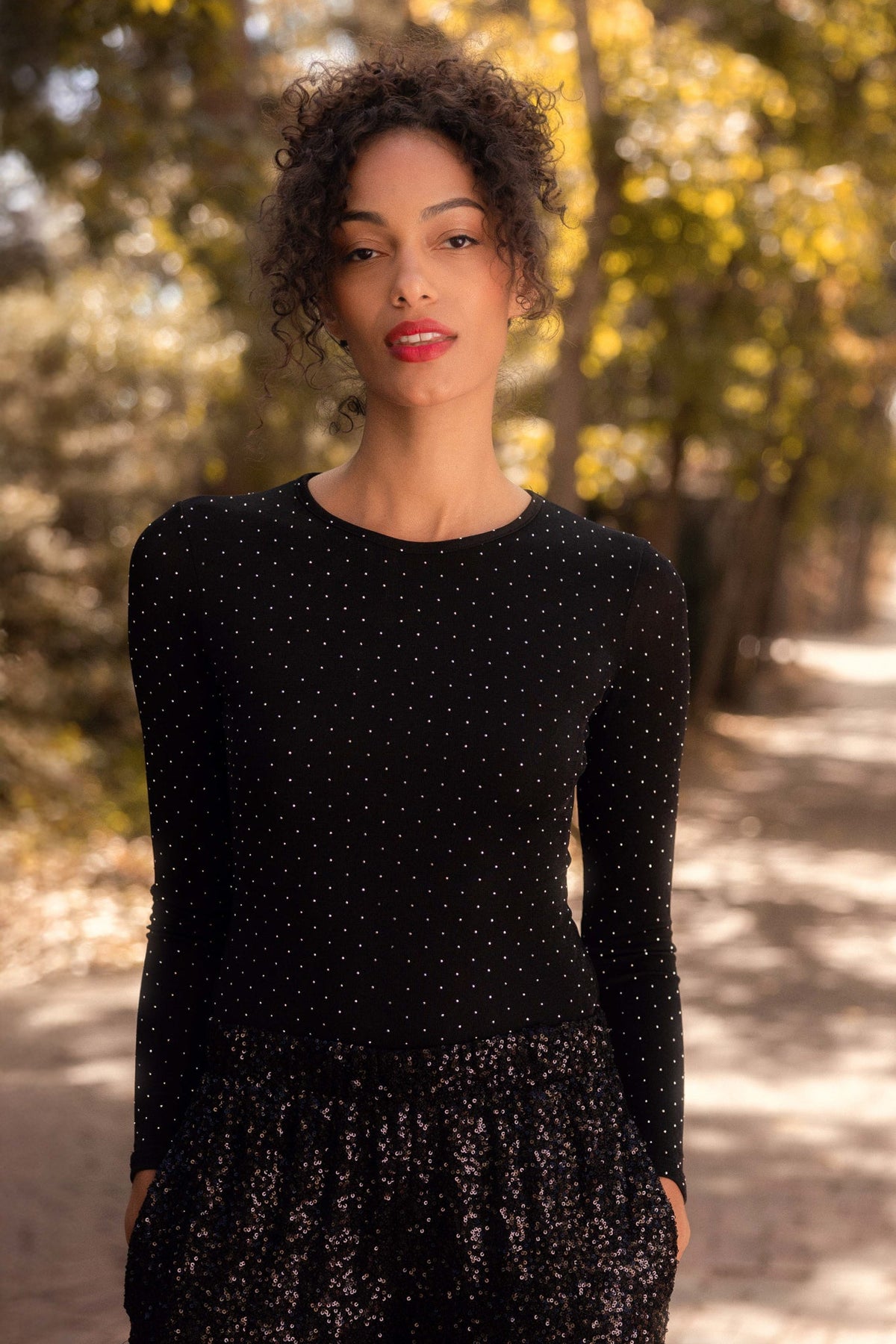 A woman with curly hair stands outdoors on a sunlit path, wearing the Bravo Top in Black from Ivy City Co—a chic, long-sleeve dotted top that is maternity friendly. Blurred trees with yellow-green leaves fill the background as she looks calmly at the camera.