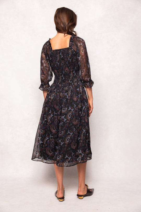 A woman stands facing away, wearing the Ivy City Co Maddison Dress in Enchanted Floral—a black, mid-length floral dress featuring sheer sleeves and ruffled cuffs. Both stylish and maternity friendly, the dress complements her brown hair styled in loose waves as she completes the look with dark open-back sandals.