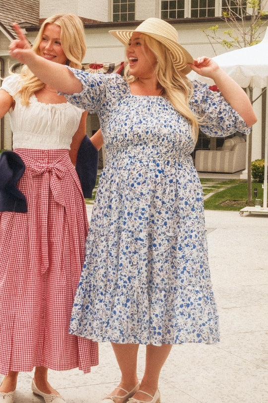 Two women stand outside, smiling. One wears the Ivy City Co Madeline Dress in Ditsy Cobalt Floral paired with a sunhat, gesturing joyfully. The other wears a white blouse and red gingham skirt with a bow. A house and tent are visible in the background.