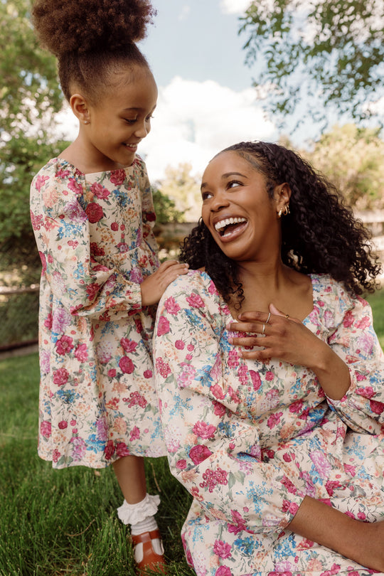 A woman and a young girl, each dressed in the Ivy City Co Mini Adelpha Dress in Autumn Amity Floral—featuring charming balloon sleeves and a smocked bodice—smile joyfully at each other while sitting and standing on grass beneath trees on a sunny day.