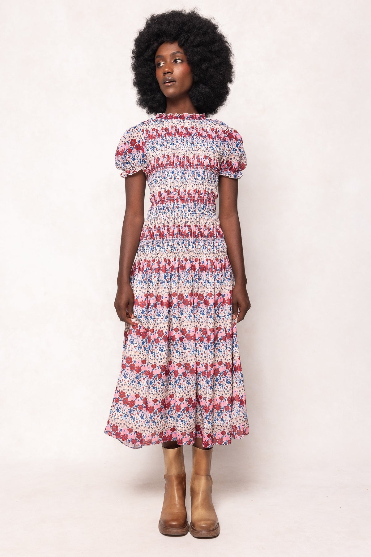 A woman with a natural afro hairstyle wears the maternity-friendly Hayden Dress in Lush Garden by Ivy City Co, a puff-sleeve, smocked floral midi dress with a red, blue, and white pattern. She stands against a plain light background and brown ankle boots.