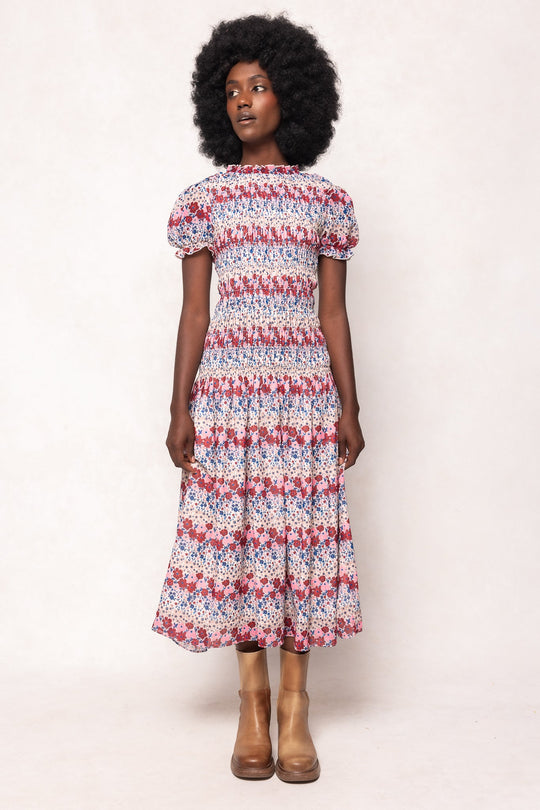 A woman with a natural afro hairstyle wears the maternity-friendly Hayden Dress in Lush Garden by Ivy City Co, a puff-sleeve, smocked floral midi dress with a red, blue, and white pattern. She stands against a plain light background and brown ankle boots.