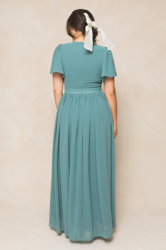 A woman stands with her back to the camera, wearing the Ivy City Co Abbie Dress in Seaglass Chiffon—a floor-length, teal gown with short sleeves. Her dark hair is styled in a low ponytail tied with a large white ribbon. Set against a light beige background, this elegant look is perfect for bridesmaids.