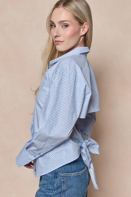 A woman with long blonde hair stands sideways against a light beige background, wearing Ivy City Co's Oriana Top in Blue—featuring blue and white stripes, a back bow detail, and paired with blue jeans.