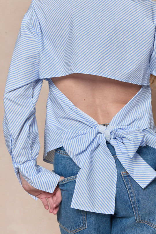 A person wears the Oriana Top in Blue by Ivy City Co—a cotton button-front shirt with blue and white stripes, open back, and a large waist bow—paired with blue jeans.