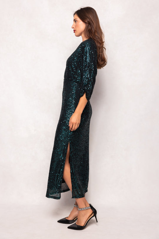 Woman wearing a long, dark green sequin dress with a slit on a white background