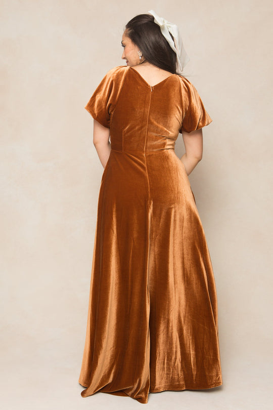 A woman with long dark hair, partially tied with a white ribbon, stands facing away to reveal the back of the Ivy City Co Tessie Dress in Amber Velvet. The nursing-friendly dress is designed with short sleeves and a V-shaped back, blending elegance and comfort.
