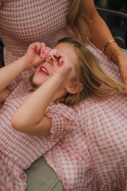 A child wearing the Ivy City Co Mini Cordelia Dress in Desert Rose Gingham playfully holds pink flowers to their eyes while laying on a woman’s lap, who is also dressed in a matching girls’ spring dress.