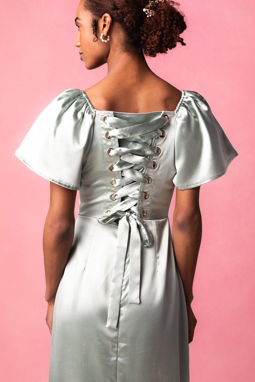 An elegant woman models the Maddie Dress in Sage Satin by Ivy City Co, featuring puff sleeves and a striking lace-up corset back. She faces away against a pink backdrop, highlighting the dress’s shiny, light green sage satin fabric.