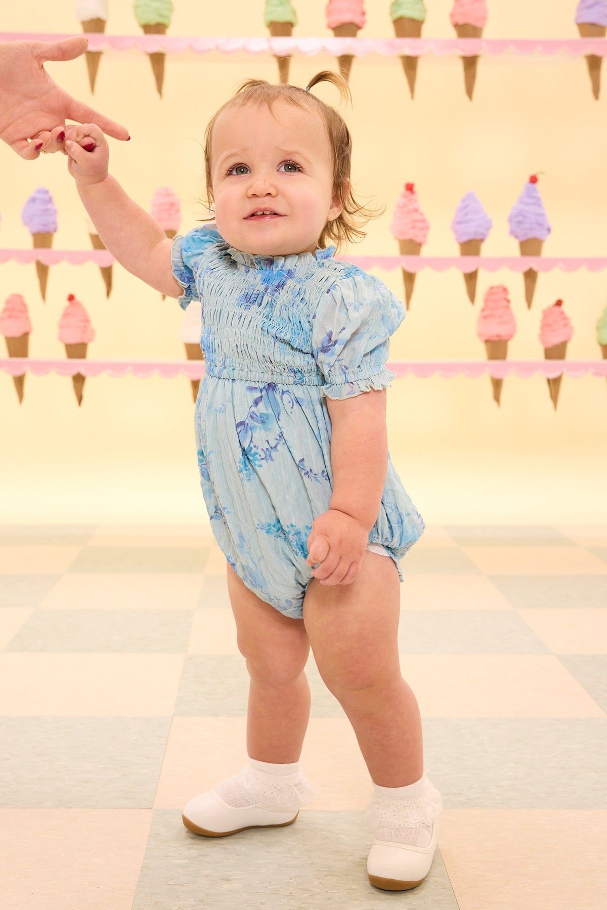 Wearing the Ivy City Co Baby Flynn Romper in Aquarelle Fleur, a toddler holds an adult’s hand while standing on a tiled floor, with pastel-colored walls adorned with ice cream cone decorations in the background.