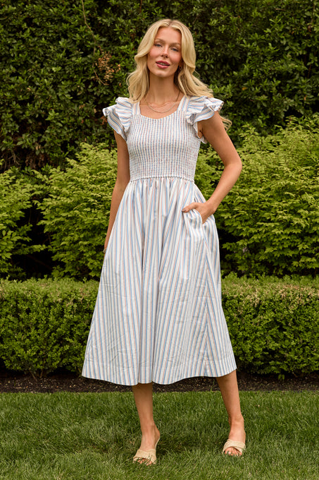 A woman with long blonde hair stands on grass in front of green bushes, wearing the Ivy City Co Hattie Dress in Heritage Stripe—a nursing-friendly midi dress with blue and tan vertical stripes, ruffled sleeves, pockets, and beige sandals.
