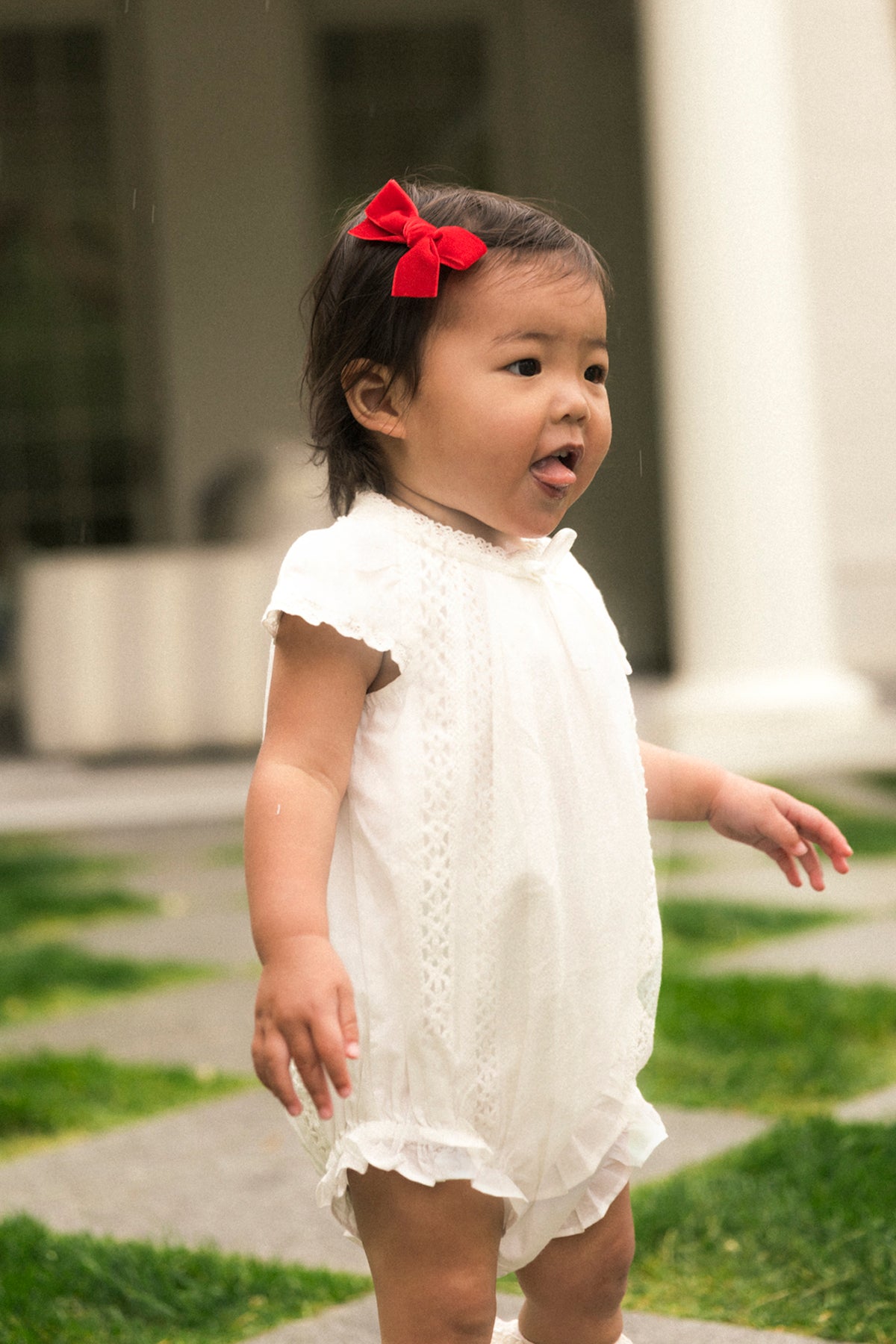 A young child playfully sticks her tongue out while standing outdoors on grass and stone, dressed in the Ivy City Co Baby Palermo Romper in White with a red hair bow.