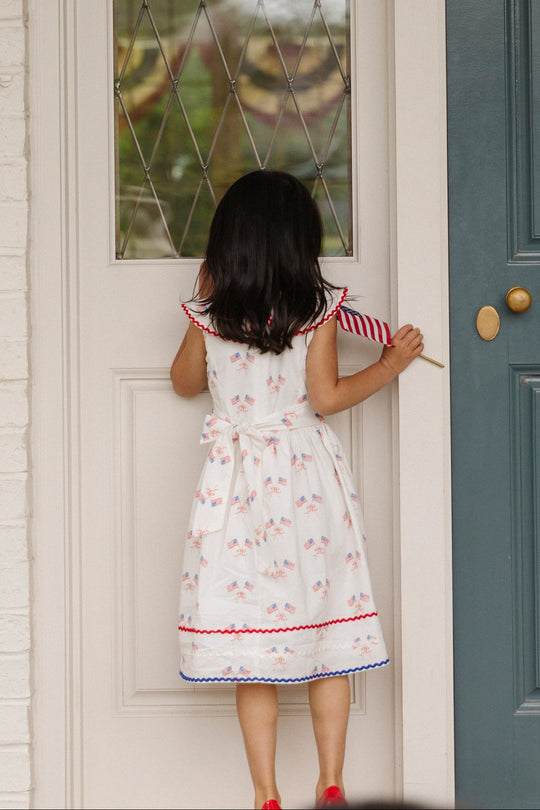 A young girl with dark hair, dressed in the Ivy City Co Mini Maisy Dress in Star Spangled, holds a small American flag and stands facing a closed front door, as if about to knock or enter.