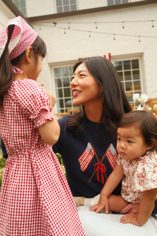 A woman wearing the Ivy City Co Freedom Sweater in Midnight smiles at a young girl in a red gingham dress and headscarf, holding a baby in a floral outfit on her lap outside near a building with large windows.
