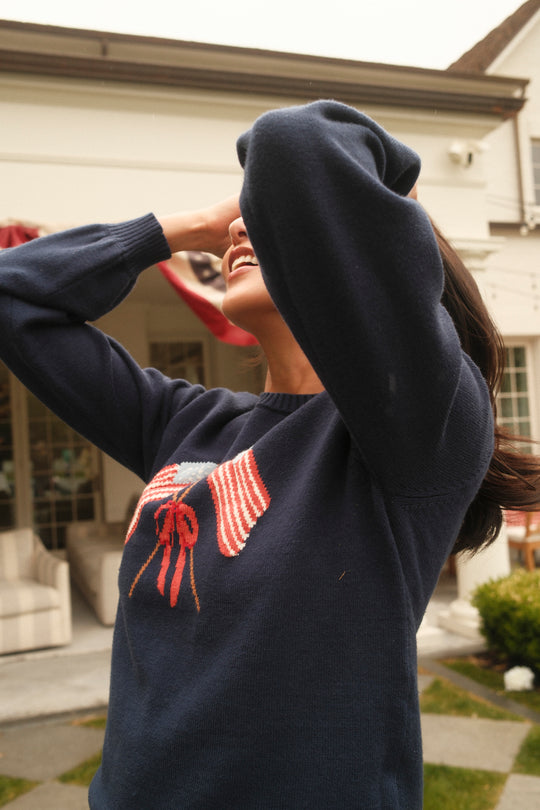 Outdoors, a smiling person covers their face with an arm and wears the Ivy City Co Freedom Sweater in Midnight, featuring two American flags and a red bow on the front. A house and greenery appear in the background.